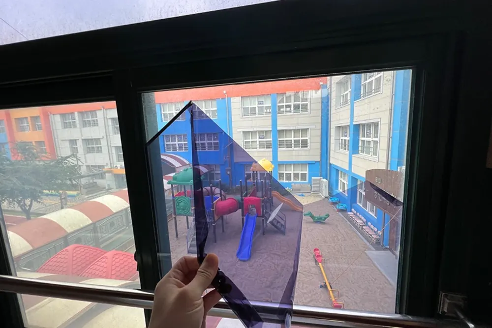 파란색 눈부심방지 필름 샘플을 창에 대어보는 모습, 창밖으로 놀이터가 보인다