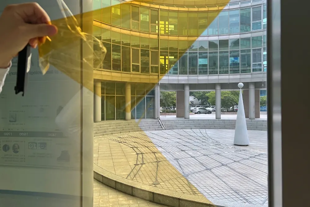 손으로 황금빛 톤의 열차단필름 샘플을 유리에 대어보는 모습